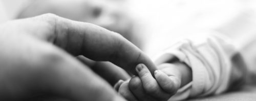 Black and white close-up of an adult's hand gently holding a baby's hand, perhaps affected by birth injuries. The soft focus emphasizes the tender interaction, highlighting the delicate fingers of the baby in contrast to the larger, protective fingers of the adult.