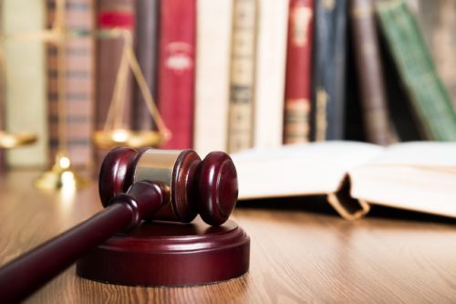 A wooden gavel rests on a sound block in the foreground. In the background, an open book on defective products and a brass balance scale sit on a wooden table, with a row of large, colorful legal books blurred in the distance, reminiscent of Washington D.C.'s legal environment.