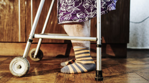 A person wearing striped socks and a floral-patterned purple dress is using a walker on a wooden floor in what appears to be a nursing home setting. The focus is on the legs and walker, hinting at movement or support, while subtly raising awareness about potential nursing home abuse.
