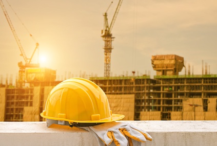 A yellow construction helmet and gloves rest on a wall at a bustling D.C. construction site. Cranes and partially built structures silhouette against the setting sun, reminding workers of the importance of safety to prevent construction accidents.