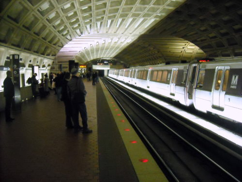 A subway train at an underground station in Washington D.C. features an illuminated arched ceiling. Passengers stand on the platform, waiting and observing. The station boasts a modern design with tiled floors and digital signs, emphasizing the efficiency of the city's mass transit system.