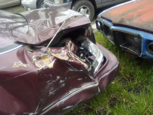 Close-up of a maroon car with significant front-end damage, likely from common truck accidents, showing a dented and crushed hood and bumper. A rusted blue car is parked beside it. Both vehicles are stationary on a grassy area.