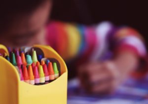 A close-up of colorful crayons in a yellow holder, with a blurred background of a child wearing a striped sweater coloring at a table in Washington D.C. daycare.