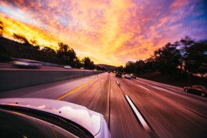A car speeds down the highway at sunset, its journey echoing the urgency of 2020. The sky is ablaze with vibrant orange and pink hues, while blurred trees and vehicles accentuate the scene's motion. From the car's perspective, this fleeting moment captures both beauty and a hint of risk.