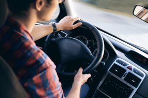 A person wearing a plaid shirt is driving a car through Washington D.C., focused on the road ahead, aware of what you need to know about avoiding car accidents. The driver's hands are on the steering wheel, with the speedometer and controls in view inside the neutral-toned interior.