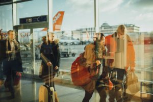 People with luggage walk through a bustling Maryland airport terminal. Sunlight streams through large windows, casting reflections on the glass. Outside on the runway, airplanes and ground support vehicles are visible, reminding travelers of what they need to know for a safe journey.
