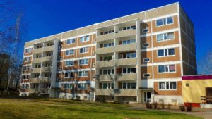 A five-story apartment building with a textured facade, multiple balconies, and white-trimmed windows under a clear blue sky. The surrounding area features grass and bare trees, hinting at winter or early spring. Tenants are advised to seek legal advice regarding potential injuries on the premises.