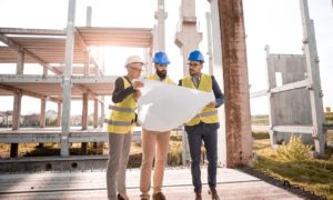 Three construction workers in safety vests and helmets examine blueprints at a bustling Washington D.C. building site. They stand on a concrete floor of an unfinished structure, surrounded by steel beams and columns, with sunlight streaming in the background, focused on what to know for their next steps.