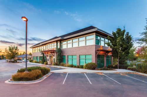A modern two-story building with large windows stands surrounded by trees and a parking lot in Washington D.C. The sky is clear with a few clouds, and a streetlamp casts light over the scene, hinting at early evening or morning tranquility.