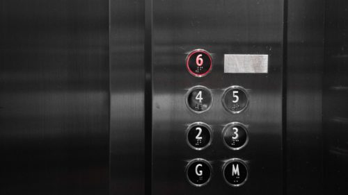 A stainless steel elevator panel showcases circular buttons for floors G, 2, 3, 4, 5, and 6. The button for floor 6 glows red while the others remain unlit. There's a rectangular plate beside the buttons that includes important information on preventing elevator accidents.