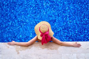 A person relaxes at the edge of a swimming pool, enjoying Memorial Day. They're wearing a straw hat with a red ribbon, and their arms are stretched out on the poolside tiles. The water reflects a vibrant blue mosaic as they unwind safely by the pool's edge.