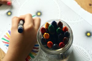 A top-down view of a child coloring a mandala with crayons at a Washington D.C. daycare. A hand holds a blue crayon, while a jar filled with various colored crayons is in the foreground. The mandala features circular and floral patterns on the paper.