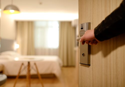 A person opens the door to a bright hotel room in Washington D.C., revealing a neatly made bed. Soft lighting and curtains create a welcoming atmosphere, ensuring comfort during your stay.