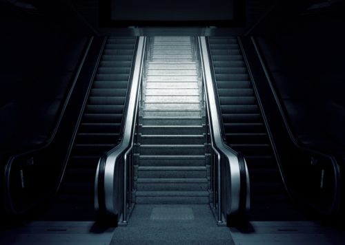 A dimly lit scene in Washington D.C. features two parallel escalators flanking a central set of stairs. The motionless escalators, casting deep shadows, evoke a mysterious and moody atmosphere while reminding us of the importance of safety to prevent escalator accidents.
