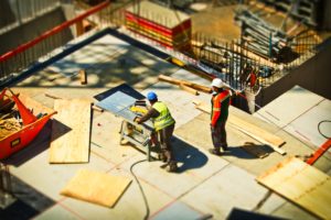 Two construction workers on a bustling construction site focus on safety as one uses a saw on plywood while the other stands nearby, ensuring accident response procedures. Both wear helmets and safety vests amidst scattered materials and tools, all framed by a low barrier.