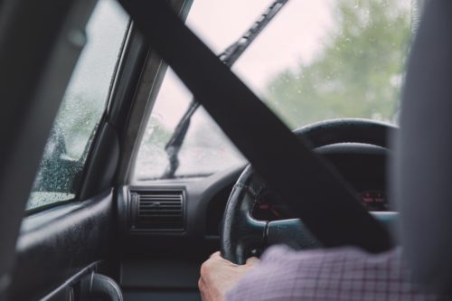 A person driving in the rain through the slick streets of Washington D.C., with a visible seatbelt and windshield wipers in motion. The road is barely visible through the wet glass, but they keep their hands securely on the slightly worn steering wheel, mindful of avoiding potential accidents.