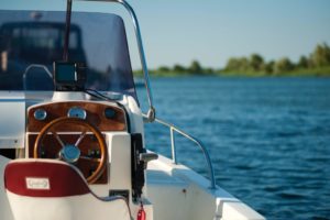 A small motorboat with a wooden steering wheel glides across a calm blue lake, its digital display shining brightly. Under a clear sky, trees embrace the distant shore—a perfect day for boating without worry of accidents or the ensuing need for compensation.