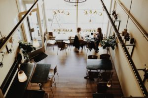 A cozy café interior with a high ceiling, featuring two people sitting at a table near the window. The café, nestled in Washington D.C., has wooden floors, several tables, and decorative plants, creating a warm atmosphere as sunlight filters through the large windows.