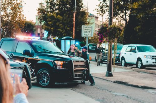 A woman is being apprehended by a police officer next to a police SUV on a Washington D.C. street. Another officer observes nearby. The scene is captured in the early evening with cars and a "Keep Right" sign in the background, raising concerns about potential police brutality.