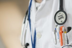 Close-up of a doctor's white coat with a stethoscope draped around the neck. The coat pocket holds several pens in blue, orange, and black. The focus is on the medical tools and pens, essential for addressing claims and ensuring patient care is free from medical malpractice.