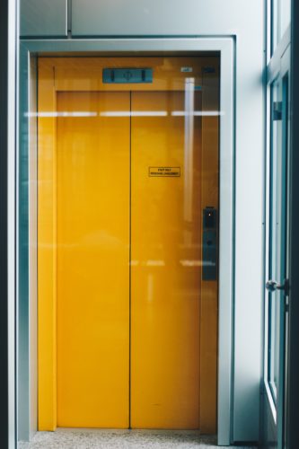 A closed yellow elevator door in a modern building in Washington D.C. features a small sign above and a control panel on the right. The light gray walls and speckled floor hint at the urbane sophistication of the capital, even as safety and proper function remain paramount to avoid any elevator accident.
