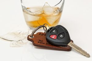 A car key with a remote on a leather keychain sits next to a shattered glass containing a brown liquid, possibly whiskey, on a white surface—a somber reminder of the dangers posed by drunk drivers.