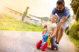 A man joyfully pushes a young child on a colorful toy tricycle along a sunlit path. The child, wearing a yellow shirt, smiles with excitement. Surrounding greenery and chalk drawings on the path enhance the playful atmosphere, offering a stark contrast to stories of daycare neglect that some families work through.