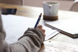 A person wearing a gray sweater is writing with a pen on sheets of paper at a wooden table, perhaps gathering notes on nursing home negligence. A white coffee cup and notebook sit nearby, against a blurred background that enhances the cozy and focused atmosphere.