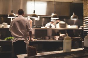 In a bustling Washington D.C. kitchen, chefs in crisp white uniforms work diligently. One chef focuses intently at a counter, while others manage plates near the stove. Overhead lights illuminate the stainless steel surfaces and shelves stacked with plates—a scene where restaurant accidents are rare but possible.