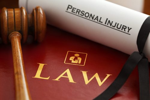 A wooden gavel rests on a red book titled "LAW" with a symbol of a scale. A rolled document labeled "PERSONAL INJURY" and mentions of insurance companies are partially visible, symbolizing legal matters related to personal injury and accident claims.