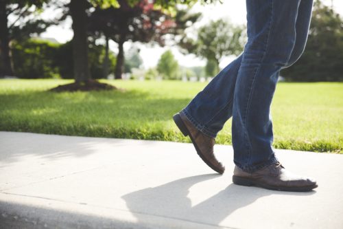 A person in jeans and brown shoes strolls along a sidewalk, with green grass, trees, and a sunny sky forming the perfect backdrop. Only their lower half is visible—a peaceful scene that contrasts sharply with the bustling concerns of Washington D.C., like pedestrian accident compensation.