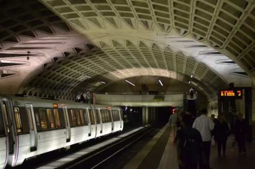 A subway station in Washington D.C. features a train stopped at the platform beneath its arched, geometric ceiling with overhead lights. People walk along the platform while an electronic sign displays train schedule information.