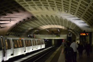 At an underground metro station in Washington D.C., a train is halted beneath an arched, geometric-patterned ceiling. Passengers stroll along the dimly lit platform as electronic signs display vital information—what you need to know for a smooth journey.