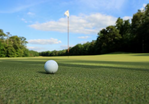 A close-up of a golf ball resting on a lush green field, with a flagstick and hole in the distance. The sky is blue with scattered clouds, and trees surround the course, highlighting the importance of golf safety as players navigate this serene landscape in Washington D.C.