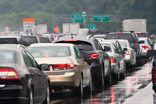 Traffic jam on a wet highway with multiple cars and a 45 mph speed limit sign. Vehicles, possibly due to a car accident, have their lights on, reflecting off the slick road. In the background, green highway signs are visible amidst the trees.