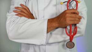 A person wearing a white lab coat stands with arms crossed, holding a red stethoscope, highlighting the importance of medical expertise. The background is a plain, neutral color.
