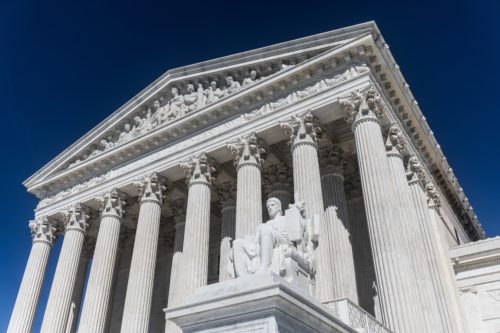 The image captures the neoclassical facade of the United States Supreme Court building in Washington D.C., with its grand columns. A statue is prominently visible in the foreground under a clear blue sky.