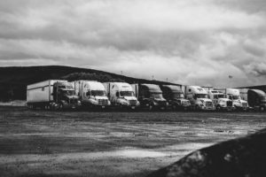 A row of parked semi-trucks in a deserted, wet parking lot under a cloudy sky captures the stark industrial setting. The black and white photo evokes an atmosphere that might follow a D.C. truck accident, with distant hills silently witnessing the scene.