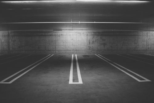 A black and white photo of an empty underground parking garage, owned by a meticulous property owner. The concrete floor has marked spaces, while walls and ceilings mirror the stark concrete backdrop. Fluorescent lights illuminate, casting shadows on the ground.