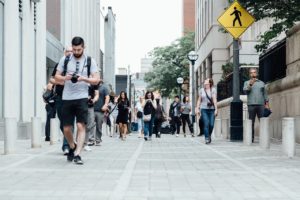 A bustling city street in Washington D.C. sees people walking in various directions, some with cameras and backpacks. A prominent pedestrian crossing sign emphasizes safety, while buildings line the street and trees loom in the background.
