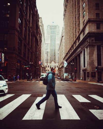A person with a backpack crosses a city street at a crosswalk in Washington D.C. The tall buildings, adorned with American flags, reflect the capital's iconic urban charm. Under cloudy skies, it's crucial to know about pedestrian accidents and stay alert in such bustling environments.