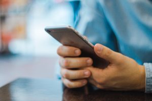 A person in a blue shirt holds a smartphone with both hands, sitting at a wooden table. The phone's side buttons are visible as they likely scroll through news or updates about the recent Washington D.C. accident. The background is softly blurred, focusing on their intent gaze.