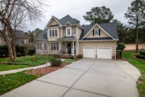 A two-story house in Washington D.C. with beige siding and a dark gray roof features a spacious driveway leading to a double garage. The front yard is landscaped with grass, trees, and shrubs, creating a welcoming suburban atmosphere, perfect for ensuring Airbnb safety during your stay.