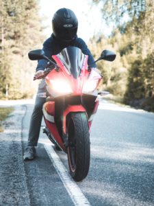 A person wearing a black helmet is sitting on a red motorcycle on a road surrounded by trees. The bike's headlights are on, emphasizing the importance of visibility as one of the key safety tips. The rider is dressed in casual attire, ready for a smooth journey ahead.