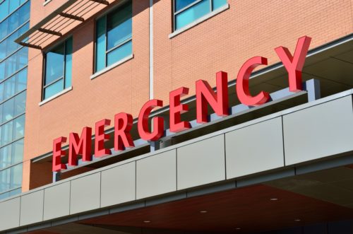 View of a hospital entrance with a large red "EMERGENCY" sign above the doorway. The building has brick walls and large glass windows, where spring rains can create slip and fall hazards on the sleek surfaces.