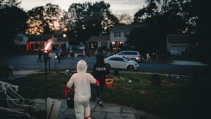 Children in Halloween costumes wander down a dimly lit street, carrying buckets for candy under the watchful Halloween moon. Houses are festively decorated, with a few kids visible in the background. One child is dressed as a ghost, another as a police officer—each ready to protect themselves from spooky surprises.