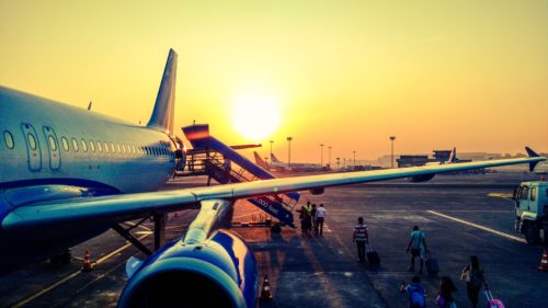 A passenger airplane is parked on the tarmac during sunset at Washington D.C. airport. People with luggage walk towards a staircase leading to the plane. The sky is a gradient of orange and yellow, casting a warm glow over the bustling scene.