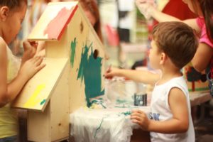 Two children enthusiastically paint a large wooden birdhouse; one applies green paint with a brush while the other dabs on splashes of yellow and red. The table is safely covered with plastic, surrounded by various painting supplies, ensuring a fun and creative experience without neglecting cleanliness.