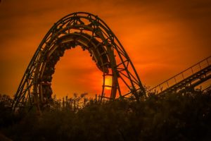 A roller coaster loop in Washington D.C. is silhouetted against a vibrant orange sunset, with the sun visible through the loop. The track is surrounded by dark foliage in the foreground, evoking memories of past amusement park thrills and the ever-important emphasis on safety.