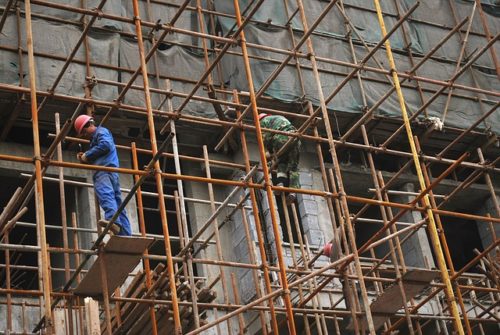 Construction workers on metal scaffolding are diligently working on the building's exterior in Washington D.C. They're wearing safety gear and helmets, surrounded by a network of poles. The facade is partly covered with green tarps, reminding all about the importance of injury compensation awareness.